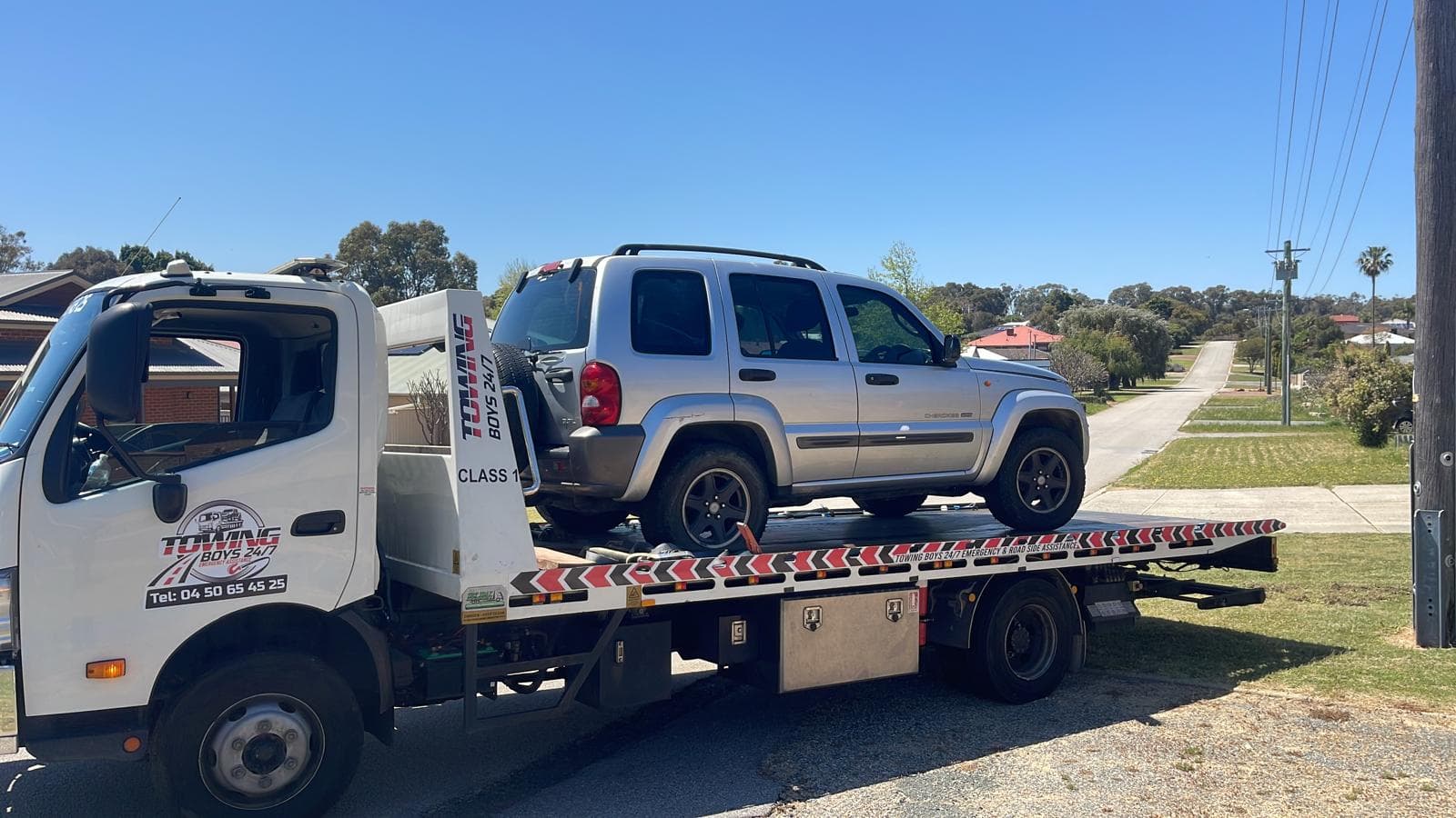 Flatbed tow truck transporting a silver Jeep Cherokee on a residential Perth street